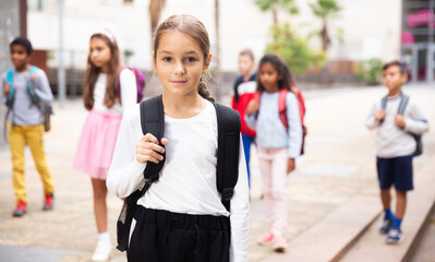 Wall Mural - Portrait of positive schoolgirl standing near school, children on background