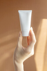 Wall Mural - Beautiful female hand holding mock-up of white plastic cream tube in rays of sunlight. Container for professional cosmetics. Concept of skin care. Close-up, brown background. Vertical image