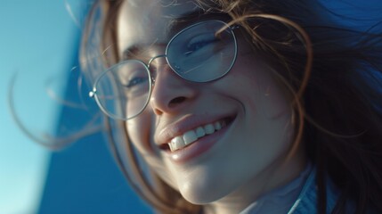 Wall Mural - A young woman with glasses smiling with her hair blowing in the wind against a blue sky backdrop.