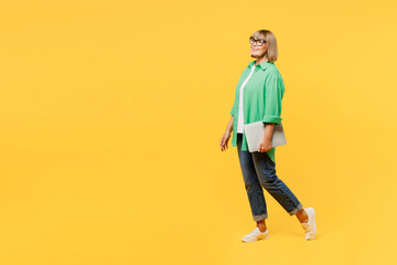 Wall Mural - Full body side profile view elderly blonde woman 50s year old wear green shirt glasses casual clothes hold use work on laptop pc computer isolated on plain yellow background studio. Lifestyle concept