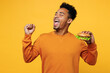 Young fun overjoyed man he wear orange sweatshirt casual clothes eat classic burger do winner gesture isolated on plain yellow background. Proper nutrition healthy fast food unhealthy choice concept.