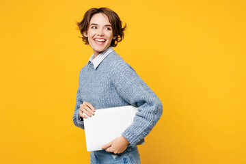 Side view young programmer IT woman she wearing grey knitted sweater shirt casual clothes hold closed laptop pc computer chatting online isolated on plain yellow background studio. Lifestyle concept.