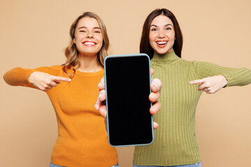 Wall Mural - Young friend two women wear orange green shirt casual clothes together hold in hand use point finger on mobile cell phone with blank screen area isolated on plain beige background. Lifestyle concept.