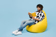© ViDi Studio - Full body smiling happy young Indian man he wears shirt white t-shirt casual clothes sit in bag chair read book isolated on plain pastel light blue cyan background studio portrait. Lifestyle concept.