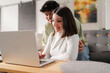 © Studio Marmellata - Cheerful young couple sitting on a sofa together, using a laptop in a relaxed home environment