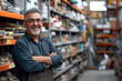 © boxstock production - Smiling middle-aged Middle Eastern man in a hardware warehouse with arms folded, standing next to a shelf containing hardware supplies