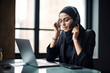 © Рика Тс - A young beautiful Muslim woman in glasses, a hijab, talks on the phone with a headset, has a pleasant conversation, looks at the computer, checks.