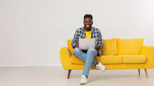 Happy young afro american man sitting in a yellow sofa and using a laptop on a white background. Networking, training, freelancing, remote work.