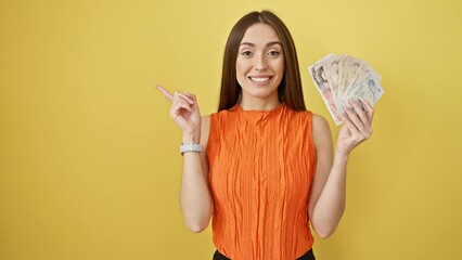 Wall Mural - Joyful hispanic beauty pointing at england pounds banknotes in her hand, smiling brightly on a yellow isolated background - a cheery indoor studio presentation