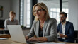 © Badi - Professional happy middle aged business woman manager explaining corporate software elearning webinar helping young employee at work looking at computer. Busy colleagues using laptop talking in office