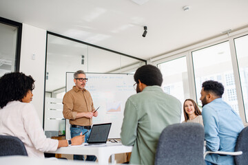 Experienced mature professional, couch or mentor, conducts a focused group discussion in a bright office, attentive team collaborates around table, reflecting a modern and efficient work environment