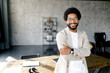 © Vadim Pastuh - A cheerful Brazilian businessman in a smart casual beige suit stands with arms crossed in loft office, his smile reflecting confidence and approachability