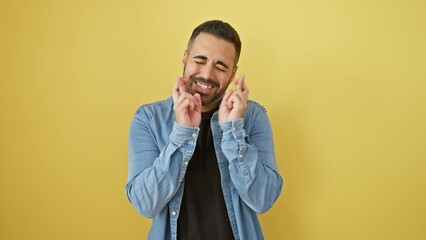 Sticker - Hopeful young hispanic man, eyes closed in wishful expression, gesturing crossed fingers for luck. stands wearing denim shirt against yellow isolated background, his superstitious symbol visible.