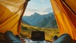 © Peeradontax - A first-person perspective from inside a tent with a laptop work remotely, and view of rugged mountain peaks under a clear blue sky.