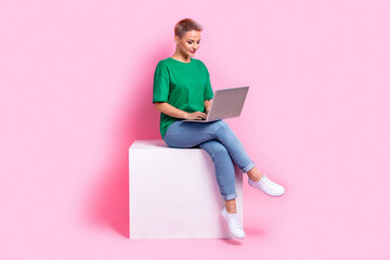 Wall Mural - Full size photo of clever girl dressed green t-shirt jeans sit on white cube typing email on laptop isolated on pink color background