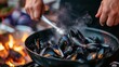 a person cooking mussels in a wok on a grill with a fire in the backgroud.