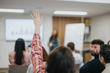 Poster - An attentive participant raises their hand to ask a question during a business seminar, with fellow attendees and presenter in the background.