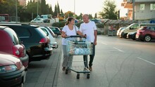 Happy Elderly Man And Woman Walking With Cart With Bottles Of Plastic Water Along Street