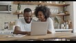 © buraratn - Pleasant family couple sitting at big wooden table in modern kitchen, looking at laptop screen. Happy young mixed race married spouse web surfing, making purchases online or booking flight tickets.