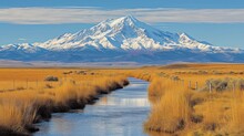 a river running through a dry grass field next to a tall snow covered mountain with a blue sky in the background.