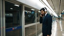 Smart Business Man Calling Phone While Wearing Headphone At Train Station. Professional Executive Manager Talking To Colleague About Marketing Strategy While Waiting For Train Or Subway. Exultant.