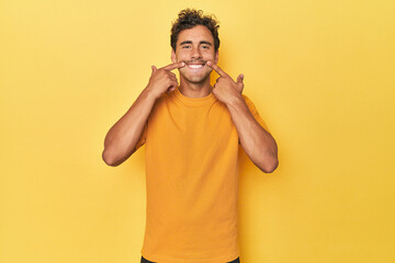 Wall Mural - Young Latino man posing on yellow background smiles, pointing fingers at mouth.