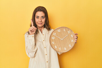 Wall Mural - Woman holding clock on yellow backdrop showing number one with finger.