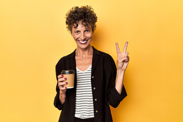 Wall Mural - Businesswoman holding takeaway coffee on yellow showing number two with fingers.