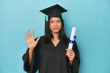 Wall Mural - Filipina graduate with diploma on blue studio smiling cheerful showing number five with fingers.