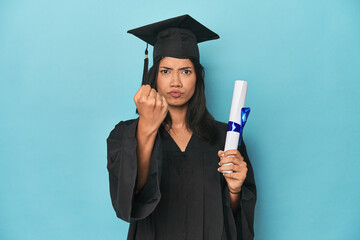 Wall Mural - Filipina graduate with diploma on blue studio showing fist to camera, aggressive facial expression.