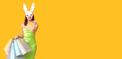 Sticker - Beautiful young woman in bunny bask holding shopping bags and credit cards on yellow background with space for text. Easter sale banner