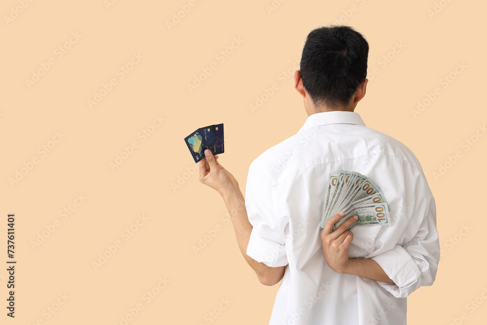 Young man with dollar banknotes and credit cards on beige background, back view