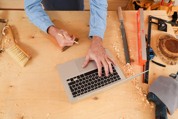 Mature carpenter working with laptop at table in shop, top view