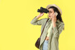 canvas print picture - Female African-American traveler with binoculars talking by mobile phone on yellow background