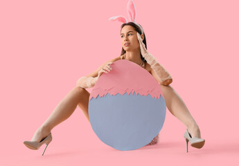 Wall Mural - Sexy young woman in bunny ears with big paper Easter egg on pink background. Easter celebration