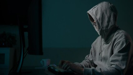 Focused unrecognizable man hacker wearing sweatshirt with hood typing on desktop computer keyboard breaking password. Dangerous hooded computer hacker infecting system with cyber virus in dark room.