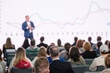© Anton Gvozdikov - Blurry audience watching a presenter at a business conference with a financial growth chart in focus.
