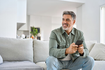 Wall Mural - Happy older mature middle aged man holding cell phone using smartphone sitting at home on sofa, relaxing on couch in living room, looking away at copy space advertising mobile offers and apps.