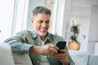© insta_photos - Smiling happy senior mature middle aged man holding cell mobile phone using smartphone sitting at home on couch, scrolling social media, checking financial apps, buying online, texting messages.