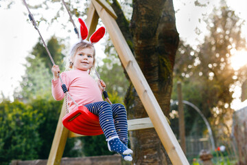 Wall Mural - Happy beautiful little toddler girl with red Easter bunny ears having fun on swing in domestic garden. Cute healthy child swinging under blooming trees on sunny spring day. Baby laughing and crying