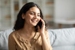 © Home-stock - Smiling indian woman talking on cellphone, enjoying pleasant mobile conversation while resting on couch at home in living room, copy space