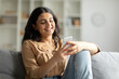 © Home-stock - Smiling hindu lady using her smartphone while relaxing on couch at home, happy woman looking online content, sitting on grey sofa in living room, copy space