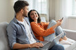 © Home-stock - Weekend pastimes. Happy indian spouses using laptop and smartphone at home, resting on sofa and looking at each other, spouses browsing internet