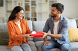 © Home-stock - Romantic gift. Loving indian man giving wrapped box to his wife, celebrating anniversary or birthday, sitting on sofa at home