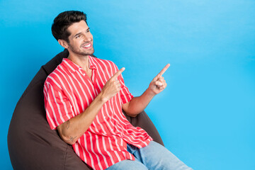 Poster - Portrait of toothy beaming guy wear stylish shirt sit on pouf look directing at sale empty space isolated on blue color background