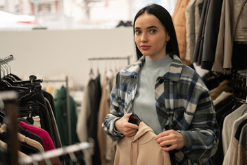 Wall Mural - A young woman makes purchases, choosing clothes at a discount in a clothing store, stands looking at the camera, thinking