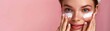 Woman Applying Moisturizing Eye Cream Close-up image of a cheerful young woman with a headband applying moisturizing cream under her eyes on a soft pink background.


