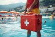 A lifeguard in yellow shorts walks along the poolside with a red first aid box, ready to provide quick medical assistance to swimmers in need