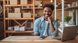 © MP Studio - young man with an afro hairstyle is smiling at the camera, and leaning on a table with a laptop in a warehouse or a small business setting.