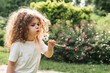 little girl blowing on dandelion with a focused expression, surrounded by greenery in a Sunlit Garden